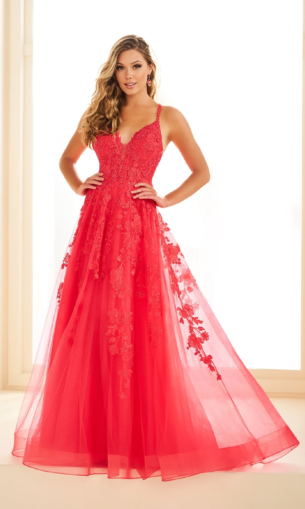 A woman stands in a bright room wearing the Ellie Wilde EW36015 long prom dress, a floor-length, sleeveless red gown with intricate lace embroidery and a fitted V-neck bodice.